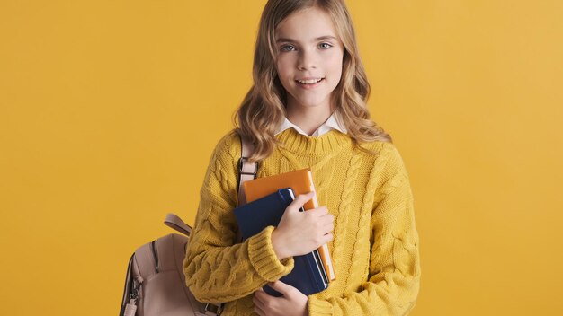 Attraente ragazza bionda studentessa vestita di maglione sorridente sulla fotocamera tenendo i quaderni e lo zaino su sfondo giallo. Concetto di studio