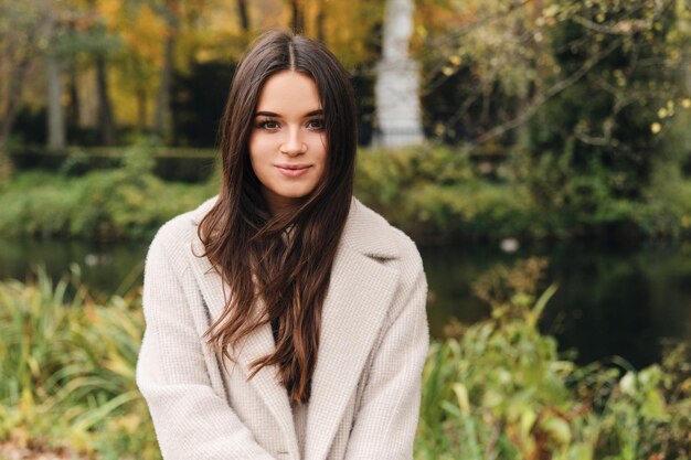 Attraente ragazza bruna in cappotto che guarda sognante a porte chiuse nel bellissimo parco cittadino