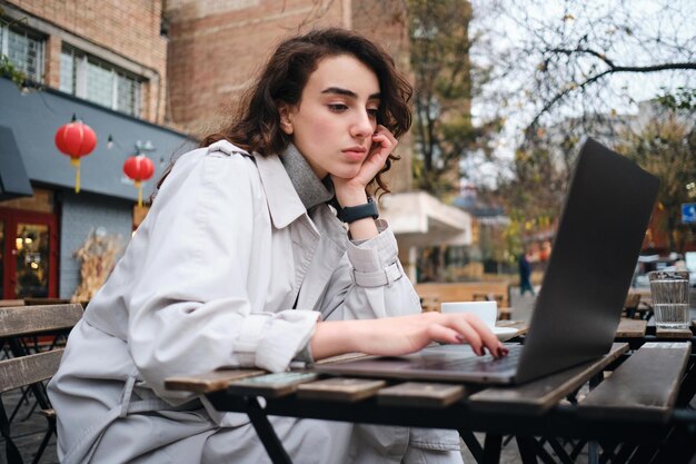 Attraente ragazza bruna seria in trench che studia sul computer portatile nella caffetteria sulla strada della città