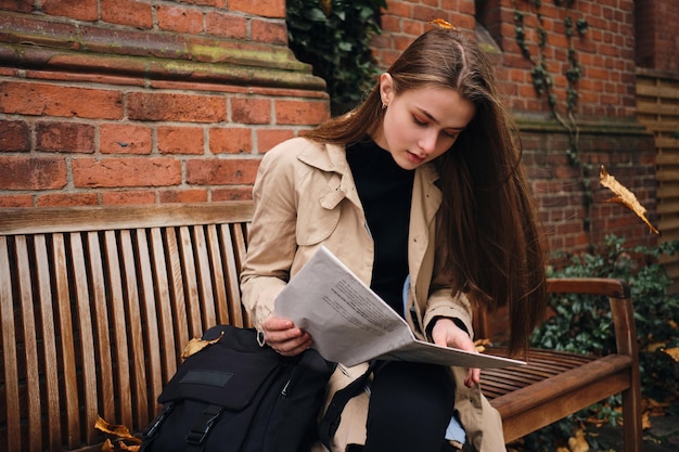 Attraente ragazza casual che legge premurosamente il giornale seduto con lo zaino sul banco all'aperto