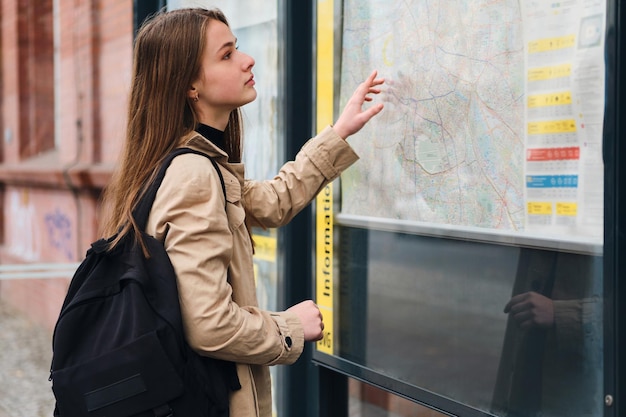 Attraente ragazza casual con zaino in cerca di mappa della città sulla fermata dell'autobus all'aperto