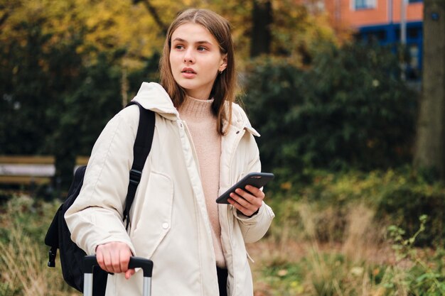 Attraente ragazza casual in piumino con zaino e cellulare che distoglie premurosamente lo sguardo all'aperto