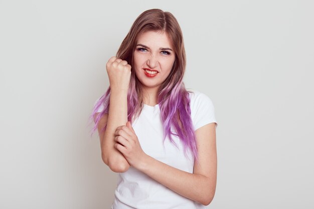 Attraente ragazza con i capelli lilla fa male al braccio, toccando la mano del dolore e guardando direttamente la telecamera, ha lesioni gravi, isolate su sfondo grigio.