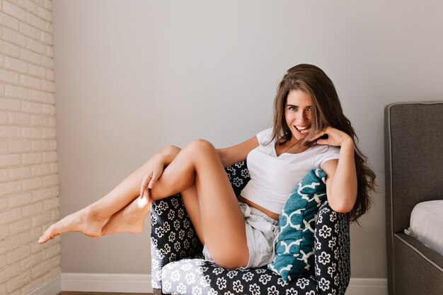 Attraente ragazza con i capelli lunghi in camicie in sedia in appartamento moderno. Lei tocca le gambe e sorride.
