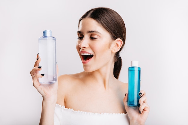 Attraente ragazza con i capelli scuri si posa sul muro bianco e sceglie che tipo di acqua micellare utilizzare.