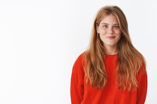 Attraente ragazza dai capelli biondi con affascinanti lentiggini e profondi occhi blu sorridente amichevole, ottimista in piedi in caldo e accogliente maglione rosso in posizione verticale essendo di buon umore in ascolto, contro il muro grigio.