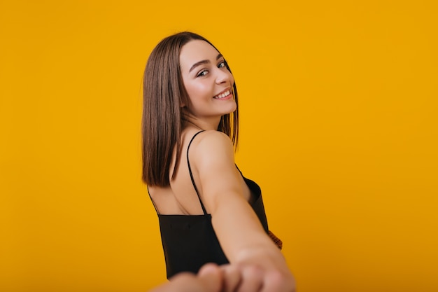 Attraente ragazza dai capelli castani con un sorriso sincero in posa. Foto dell'interno dal retro della straordinaria modella in abito nero.