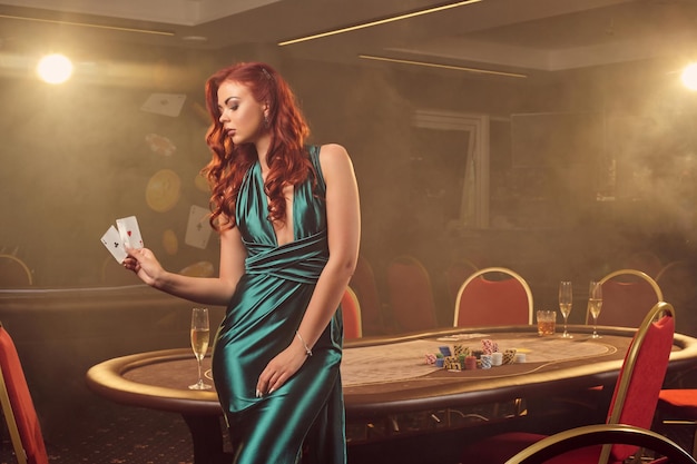 Attraente ragazza dai capelli rossi in un lungo abito di raso blu è in posa con due assi in mano contro un tavolo da poker in un casinò di lusso. Passione, carte, fiches, alcol, vincita, gioco d'azzardo - è come enterta femminile