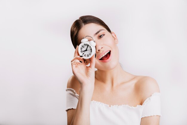 Attraente ragazza dai capelli scuri carino sorride e tiene piccolo orologio, in posa sulla parete isolata.