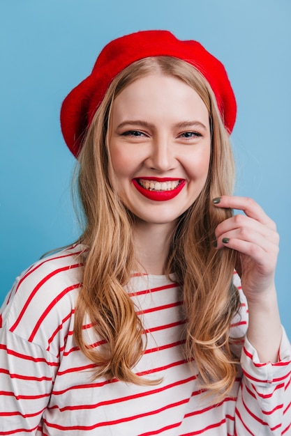 Attraente ragazza in berretto francese in posa con un sorriso sincero. donna bionda accattivante isolata sulla parete blu.