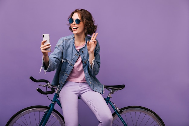 Attraente ragazza in pantaloni viola che fa selfie. Buon umore giovane signora in giacca di jeans che si siede sulla sua bicicletta.