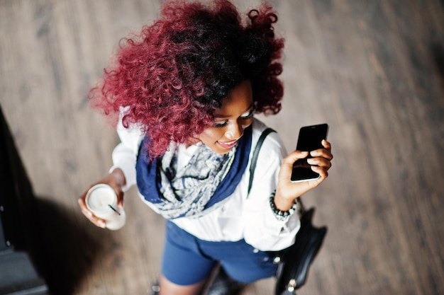 Attraente ragazza riccia afroamericana in camicetta bianca e pantaloncini blu poste al bar con latte e telefono cellulare a portata di mano Vista dall'alto