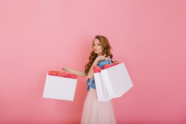 Attraente ragazza riccia in camicia di jeans che tiene grandi sacchi bianchi dal negozio di abbigliamento isolato su sfondo rosa. Affascinante giovane donna dai capelli lunghi in abito carino in posa con i pacchetti dopo lo shopping.