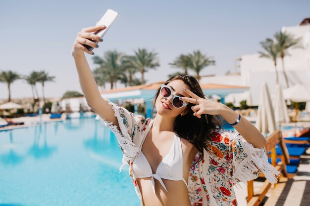 Attraente ragazza sorridente divertendosi sul resort e facendo selfie sul paesaggio meridionale con palme esotiche. Giovane signora abbronzata esile in bikini bianco che prende foto di se stessa che mostra il segno di pace