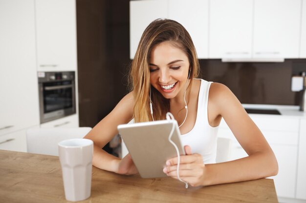 Attraente ragazza sorridente seduta a tavola e ascoltando musica controllando qualcosa in tablet