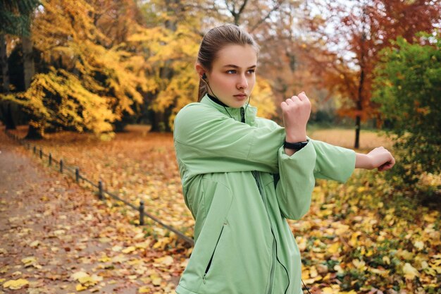 Attraente ragazza sportiva in auricolari che si allunga intensamente prima di correre nel parco cittadino d'autunno