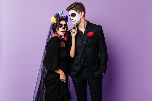 Attraente ragazza zombie in corona colorata in piedi con il fidanzato. Allegro coppia caucasica in costumi muertos in posa di halloween su sfondo viola.