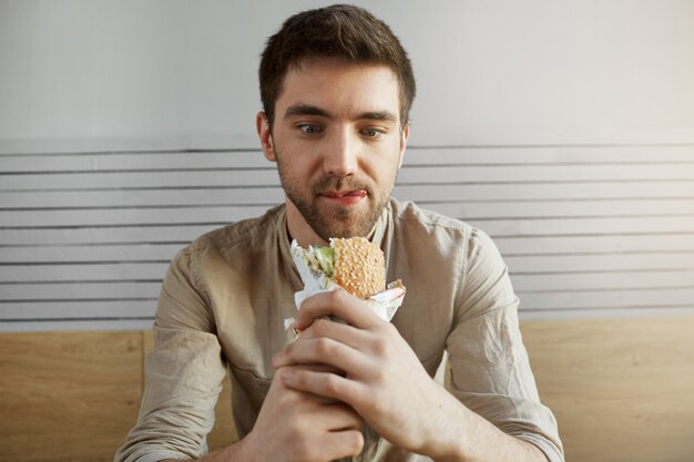 Attraente ragazzo dai capelli scuri seduto nel caffè, guardando con espressione felice a sandwich, essendo felice di mangiare qualcosa dopo tutto il giorno al lavoro. Uomo affamato che va a mangiare hamburger.