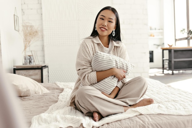Attraente signora bruna in cardigan beige e pantaloni sorride guarda nella fotocamera e abbraccia il cuscino in camera da letto Giovane donna asiatica in abito elegante si siede su un letto morbido