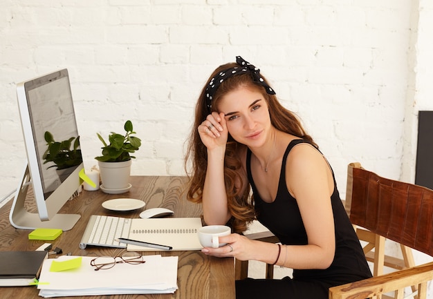 Attraente sorridente giovane femmina libero professionista gustando il cappuccino durante la pausa caffè mentre lavora da remoto nel suo ufficio a casa, utilizzando la connessione Internet ad alta velocità sul computer dello schermo dello spazio di copia