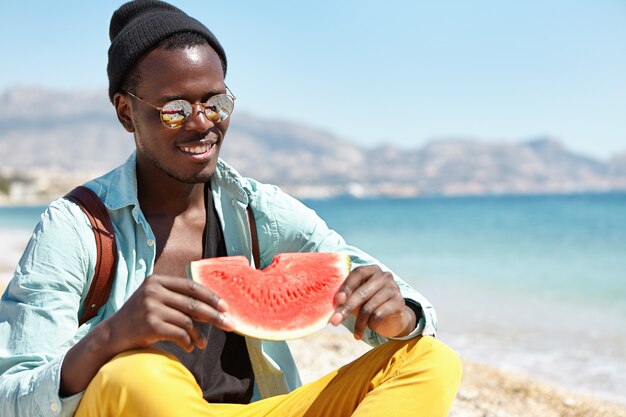 Attraente studente africano di buon umore che indossa abiti eleganti e accessori che riposano sulla spiaggia dopo il college in una giornata di sole, gioendo per il bel tempo sul mare e mangiando anguria matura