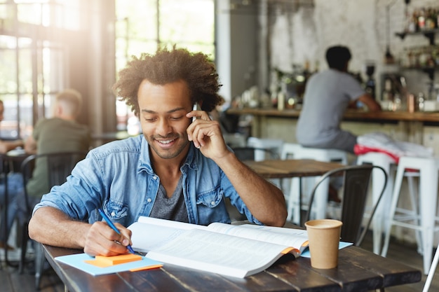 Attraente studente afroamericano facendo compiti a casa alla mensa universitaria, seduto al tavolo con un libro di testo e una tazza di caffè, prendendo appunti e parlando al cellulare, con sguardo felice