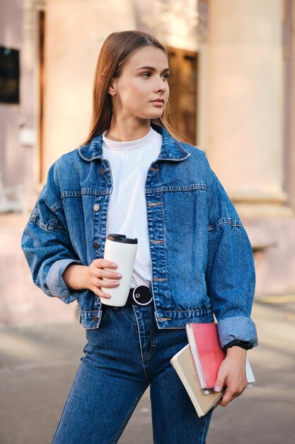 Attraente studentessa casual elegante in giacca di jeans con caffè e libri di testo che distolgono premurosamente lo sguardo all'aperto