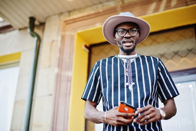 Attraente uomo afroamericano in camicia a righe cappello e occhiali da sole che cammina per strada nel centro cittadino