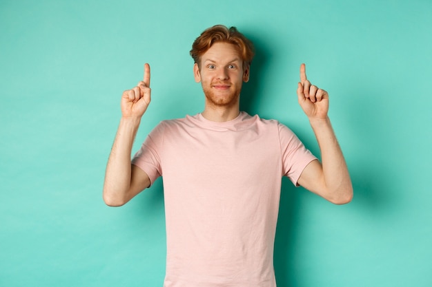 Attraente uomo barbuto con i capelli rossi, che indossa una t-shirt, sorride allegro e punta le dita in alto, mostra pubblicità, in piedi su sfondo turchese.