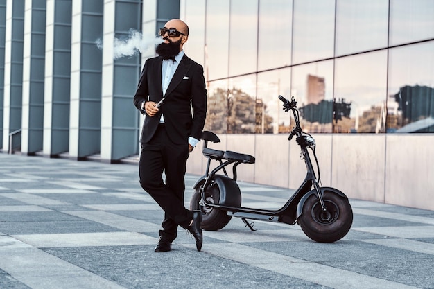 Attraente uomo calvo in occhiali da sole sta fumando, facendo un bel vapore mentre è in piedi vicino alla sua bici elettrica.