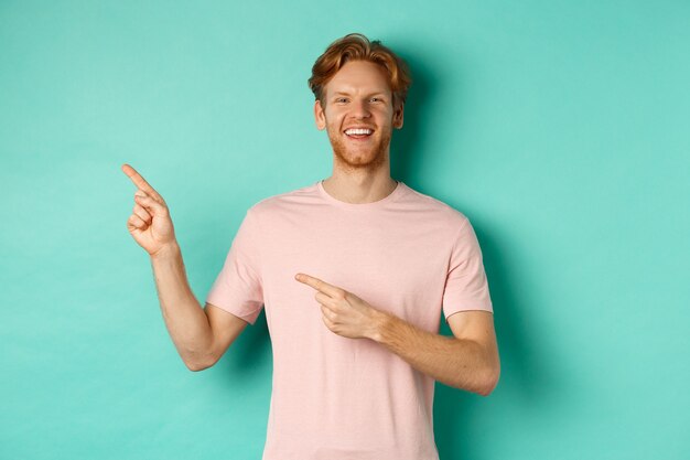 Attraente uomo caucasico in t-shirt puntando le dita a sinistra, sorridendo felice e mostrando pubblicità, in piedi su sfondo turchese. Copia spazio