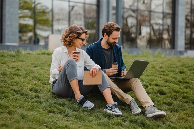 Attraente uomo e donna sorridenti che parlano seduti sull'erba nel parco urbano, prendendo appunti