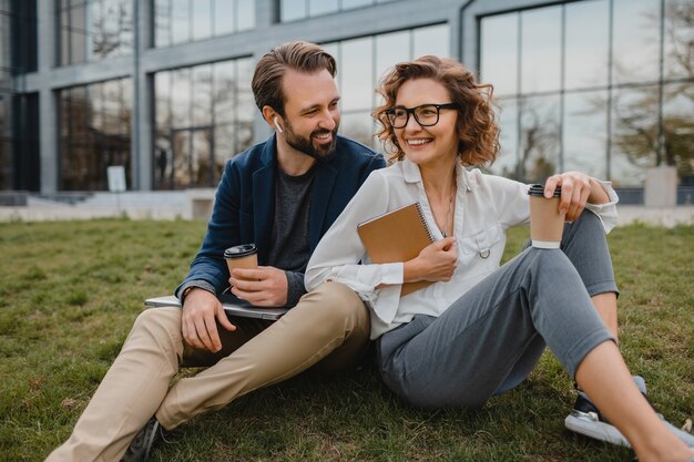 Attraente uomo e donna sorridenti che parlano seduti sull'erba nel parco urbano, prendendo appunti