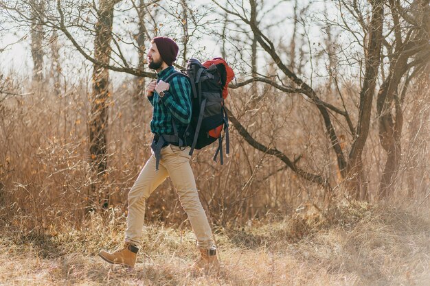 Attraente uomo hipster che viaggia con lo zaino nella foresta autunnale che indossa camicia e cappello a scacchi