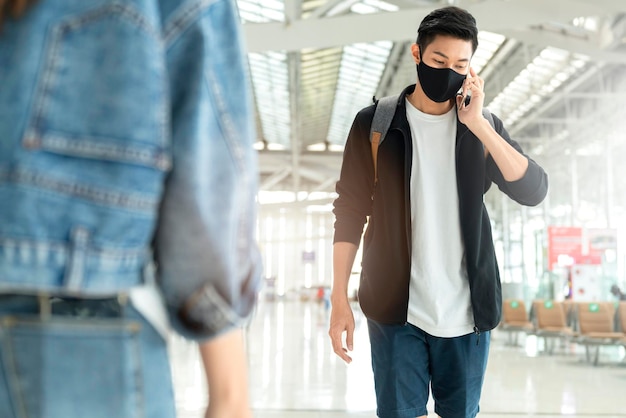 Attraente vestito casual da treveller maschio asiatico indossa una maschera protettiva nera che cammina e usa la mano per comunicare con lo smartphone nel terminal dell'aeroporto