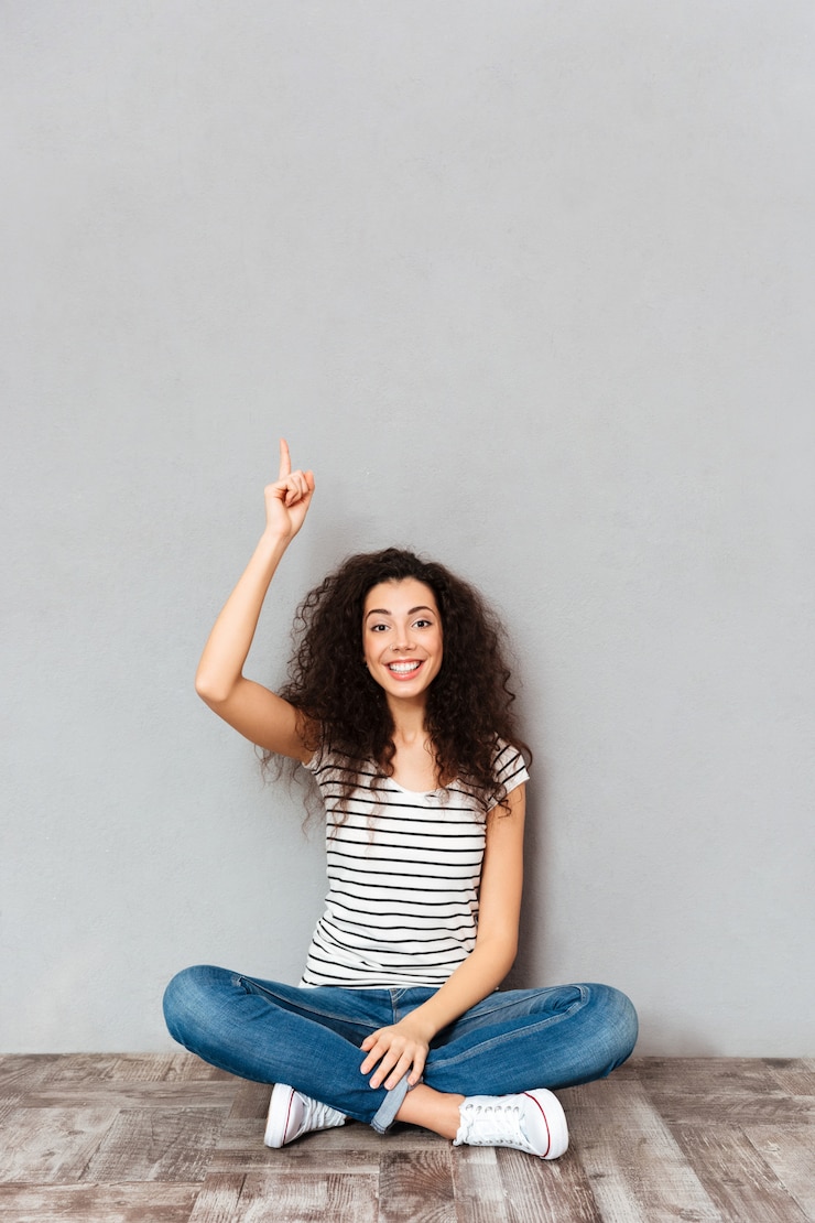 Foto gratuito avere idea! la donna sveglia con capelli scuri che si siedono con le gambe ha attraversato sul pavimento che mette il dito indice su che significa l'eureka sopra la parete grigia