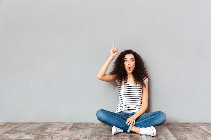 Foto gratuito avere idea! la donna sveglia in abbigliamento casual che si siede con le gambe ha attraversato sul pavimento che gesturing il dito indice su che significa eureka sopra la parete grigia