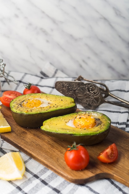 Avocado diviso a metà con uovo york; pomodori e fettina di limone sul tagliere sopra il tovagliolo con pinza antica