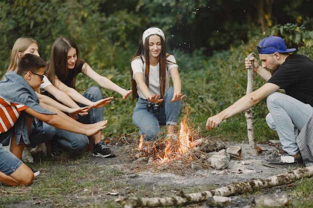 Avventura, viaggi, turismo, escursione e concetto di persone. Gruppo di amici sorridenti in una foresta. Persone sedute vicino al falò.