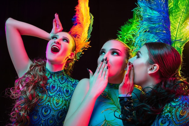 Avvicinamento. Belle giovani donne in carnevale, elegante costume in maschera con piume su sfondo nero in luce al neon.