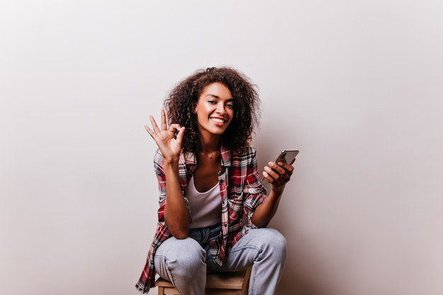 Avvincente ragazza africana in posa con il segno giusto. Ricci signora nera con un sorriso romantico che tiene smartphone.