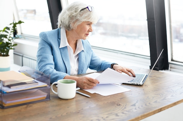 Avvocato donna anziana con esperienza di successo che indossa un bel vestito e occhiali sulla testa utilizzando un computer portatile sul posto di lavoro, guardando lo schermo con espressione facciale concentrata