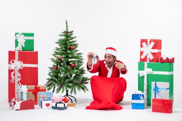 Babbo Natale che indica qualcosa seduto per terra e che mostra l'orologio vicino a regali e albero di Natale decorato