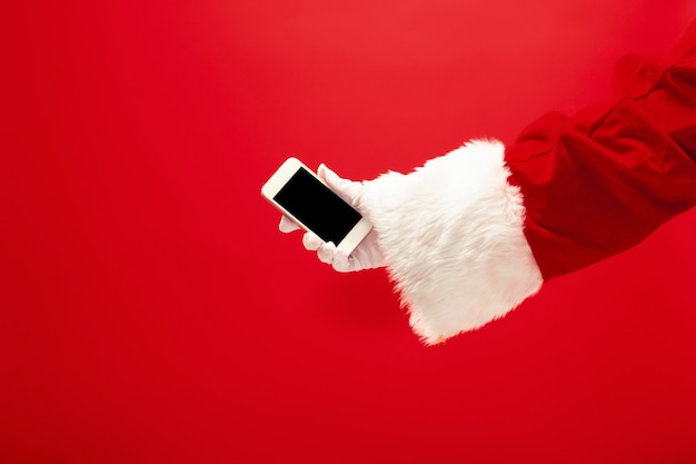 Babbo Natale tenendo il telefono cellulare pronto per il tempo di Natale su sfondo rosso studio. La stagione, l'inverno, le vacanze, la celebrazione, il concetto di regalo