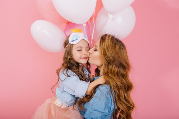 Bacio carino di giovane mamma riccia in camicia di jeans e adorabile figlia in maschera per dormire alla festa di compleanno. Piccola ragazza dai capelli lunghi in gonna lussureggiante che bacia e abbraccia sua madre, sinceramente grazie per l'evento divertente