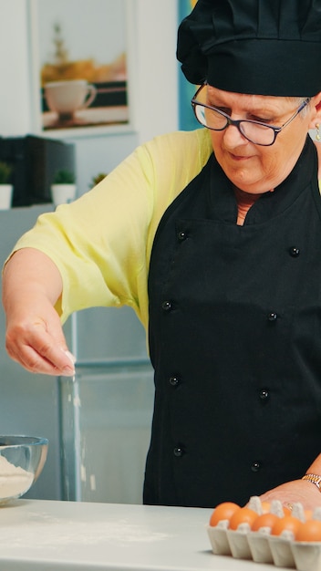 Baker spalmare la farina sul tavolo di legno a casa in cucina moderna indossando grembiule e bonete. Felice chef anziano con una spolverata uniforme, setacciando setacciando gli ingredienti grezzi a mano cuocendo la pizza fatta in casa