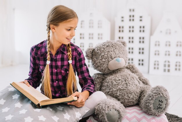 Bambina adorabile che legge un libro con il suo orsacchiotto