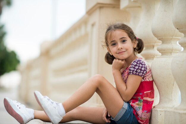 Bambina adorabile pettinata con le trecce all&#39;aperto seduto sul pavimento urbano.