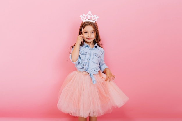 Bambina allegra con capelli lunghi del brunette in gonna di tulle che tiene la corona della principessa sulla testa isolata su fondo rosa. Celebrando il carnevale luminoso per i bambini, la festa di compleanno, il divertimento di un bambino carino