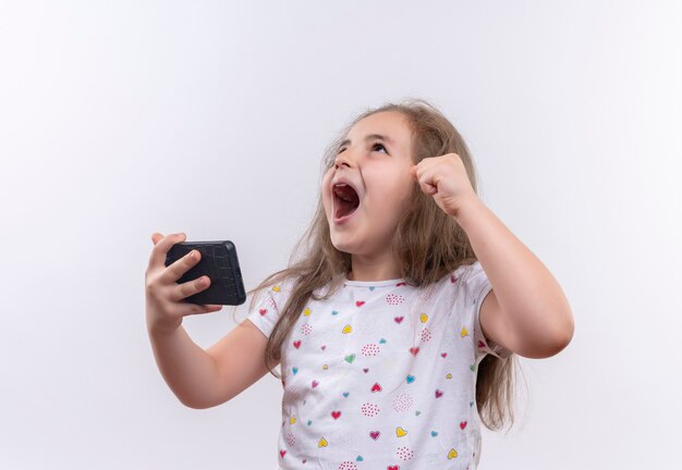 Bambina allegra della scuola che indossa il telefono bianco della tenuta della maglietta sulla parete bianca isolata